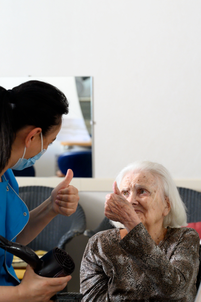 une résidente âgée lève le pouce en direction d'une soignante en tenue professionnelle